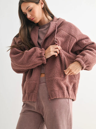 Model in mauve teddy fleece hoodie with hands in pockets and front zip closure.