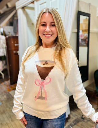 Woman smiling in a cream knit sweater with an espresso martini graphic and pink bow, standing inside a boutique.