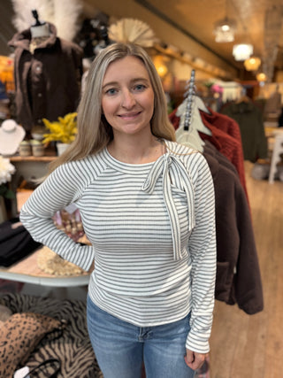 Woman smiling and wearing a fitted ivory and grey striped ribbed long-sleeve top with a bow tie detail at the neckline, paired with blue jeans inside a boutique setting.