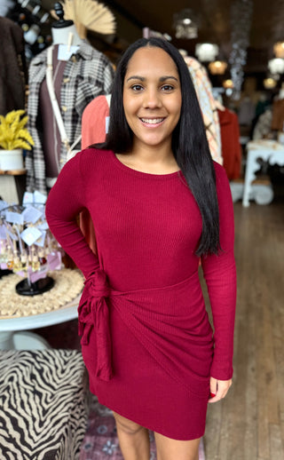 Woman wearing a burgundy ribbed knit long sleeve dress with side tie detail, standing in a boutique surrounded by fall clothing displays.