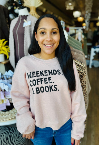Woman wearing a soft pink graphic sweatshirt that says "Weekends, Coffee, and Books" on the front of the sweatshirt.