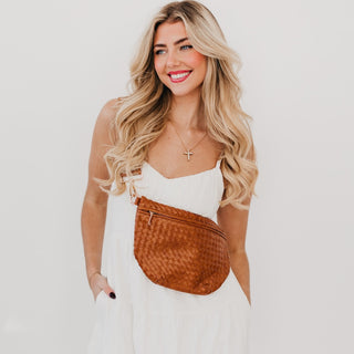 Model wearing the solid camel brown Westlyn woven bum bag with a white dress — bag styled crossbody with an adjustable strap and gold hardware.