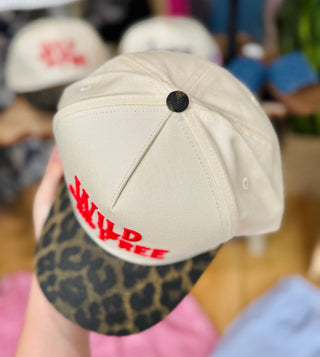 Top view of the tan trucker hat showing its structured crown, red embroidered front, black top button, and leopard print bill.