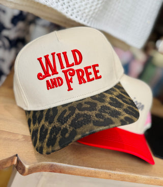 Tan trucker hat with bold red “WILD AND FREE” embroidery on the front panel and a curved leopard print bill, displayed on a wood surface.