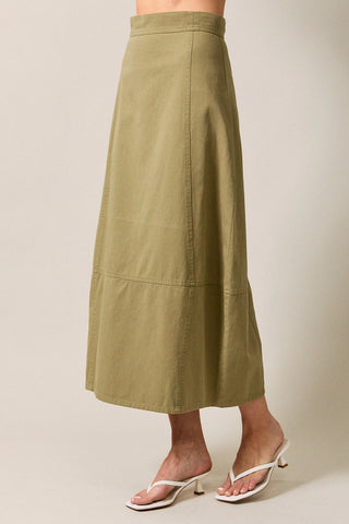 An olive green midi skirt worn by a model, paired with white heeled thong sandals, shown from the waist down against a neutral beige studio background.