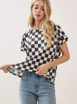 Front view of a woman wearing a black and white checkered knit short-sleeve top and blue jeans, holding the hem of the top to show its drape and stretchability against a plain light background.