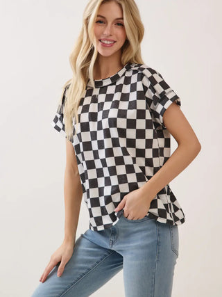 Side view of a woman modeling a black and white checkered short-sleeve top with raw edge details, paired with light blue jeans, against a plain off-white background.