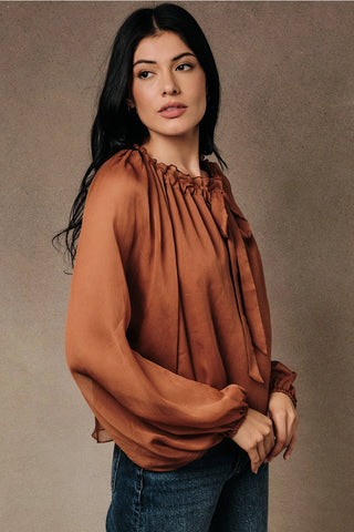 Side view of rust sheer chiffon ruffled tie neck blouse with gathered peasant silhouette and long sleeves