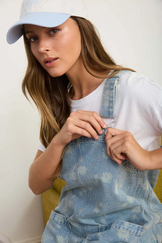 Woman wearing a light blue cap and denim overall dress against a white background. Woman is showing that straps can be adjusted and taken off from the front.
