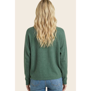 A woman viewed from the back wearing a forest green knit sweater and blue jeans against a solid light beige background.