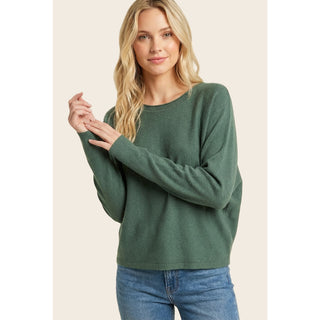 A woman wearing a forest green, crewneck, dolman, long-sleeve, knit sweater and blue jeans, posing against a solid light beige background.