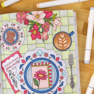 Coloring book with floral and meal-themed designs on a wooden table with markers.