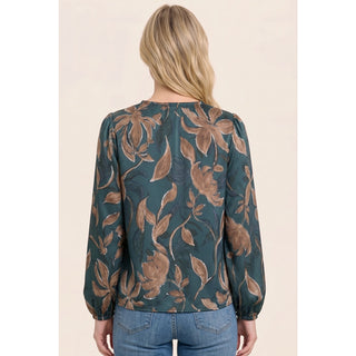 A woman viewed from the back wearing a long-sleeve blouse with a forest green and brown floral pattern, paired with blue jeans, against a solid light beige background.