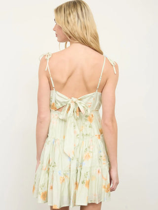Back view of a woman wearing a light green floral dress with thin straps and tie bow back detailing.