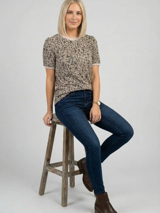 Leopard print short sleeve knit top styled casually with skinny jeans, model seated on wooden stool, relaxed fit shown tucked into jeans