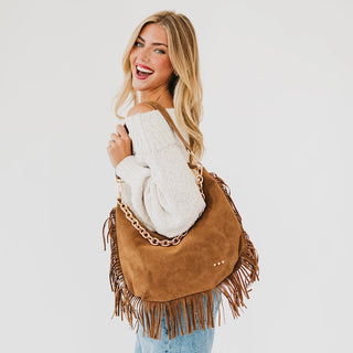 A woman wearing a white knit sweater and blue jeans carries the Loretta large camel suede shoulder bag with fringe and a gold chain strap against a light gray studio background.