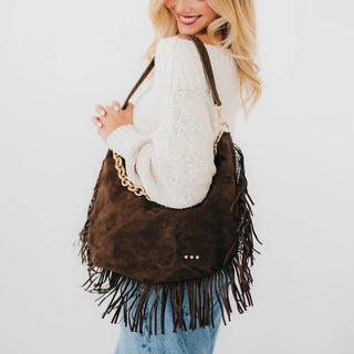 A woman wearing a white knit sweater and blue jeans carries the Loretta large brown suede fringe shoulder bag with a gold chain strap against a plain white background.