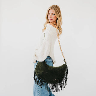 A woman wearing a white knit sweater and blue jeans carries the Loretta olive green suede crescent-shaped fringe bag with a gold chain strap against a plain white background.