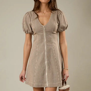 A woman wearing a cream and brown vertically striped mini flare dress with puff sleeves and a front, full zipper, and v-neck design, holding a small handbag against a plain beige background.
