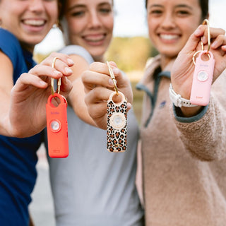 Three women holding She's Birdie personal safety keychain alarms in coral, leopard print and pink.
